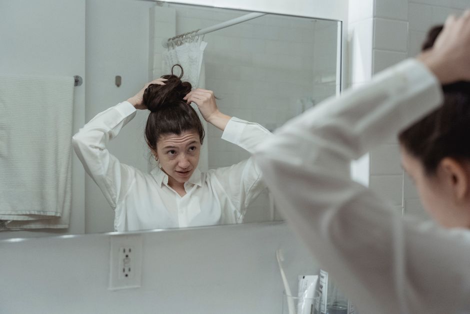 Woman styling her hair in front of bathroom mirror wearing a white shirt.
