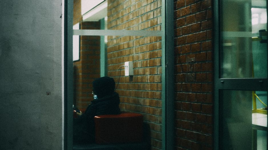 Person in a hoodie sitting indoors against a brick wall, using a phone.