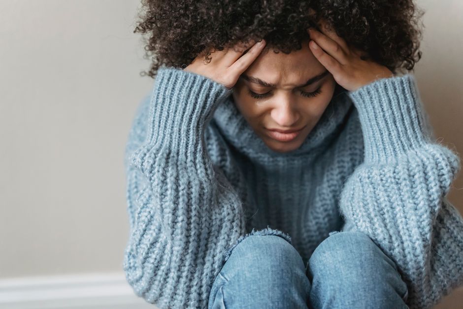 Frowning African American female with short curly hair in casual clothes sitting on floor and touching head against light wall