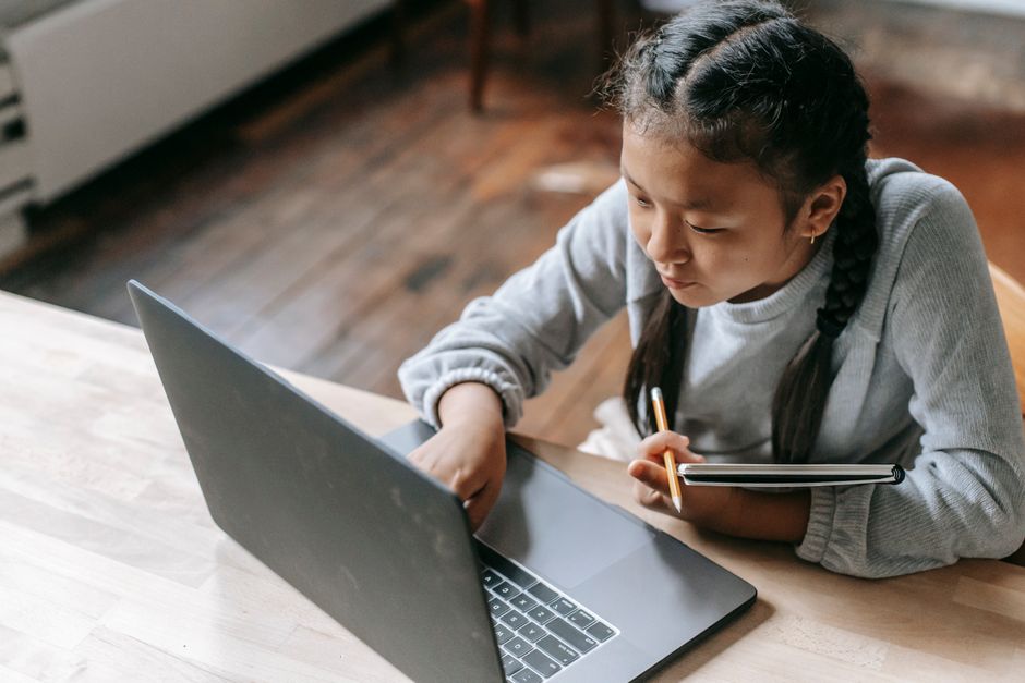From above of concentrated Asian schoolgirl with pigtails sitting at wooden table with copybook and pencil and browsing laptop for school task