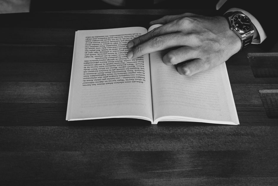 A monochrome photograph of a man's hand pointing at a book on a wooden table.