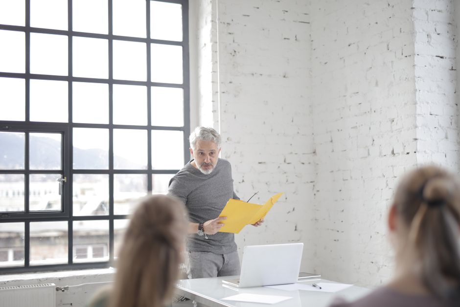 Serious aged gray haired bearded male speaker in casual wear answering question at meeting in modern office boardroom while standing against table with laptop and presenting ideas to colleagues