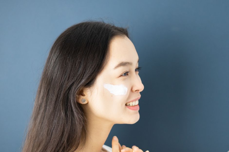 Profile of a woman with cream on her cheek against a blue background, highlighting skincare routine.