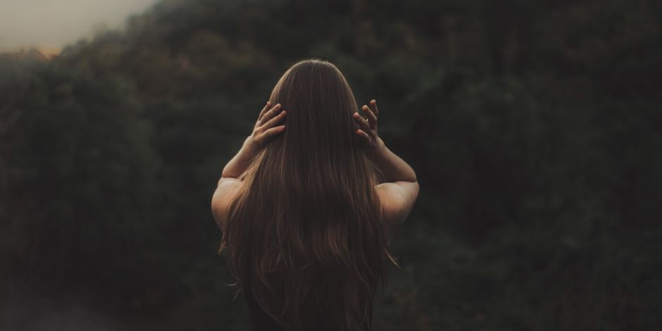 A woman with long hair stands in a forest, backlit by soft light, with her hands in her hair.