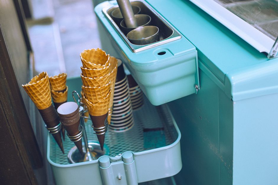 From above of light blue ice cream cart equipped with metal glasses with scoop above basket with waffle cones and paper cups